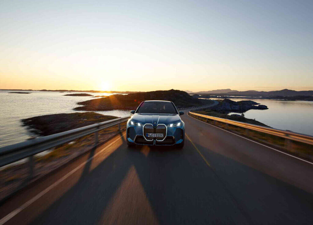 Ein blauer BMW iX fährt bei Sonnenuntergang auf einer Küstenstraße, die Sonne steht tief am Horizont, umgeben von Wasser und felsigen Inseln. - AH Praller
