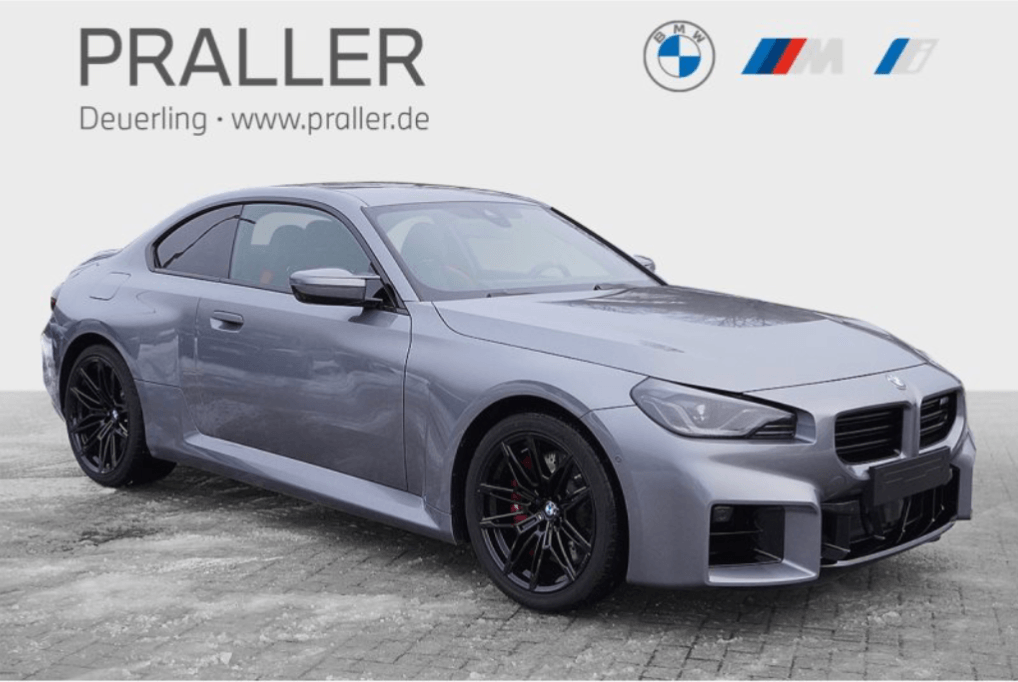 Ein silbernes BMW Coupé mit schwarzen Rädern ist auf einer Steinfläche geparkt. Im Hintergrund weisen das PRALLER-Logo und BMW M2 Plaketten auf exklusive BMW M Angebote der Autohaus Praller GmbH hin, zu sehen auf www.praller.de. - AH Praller
