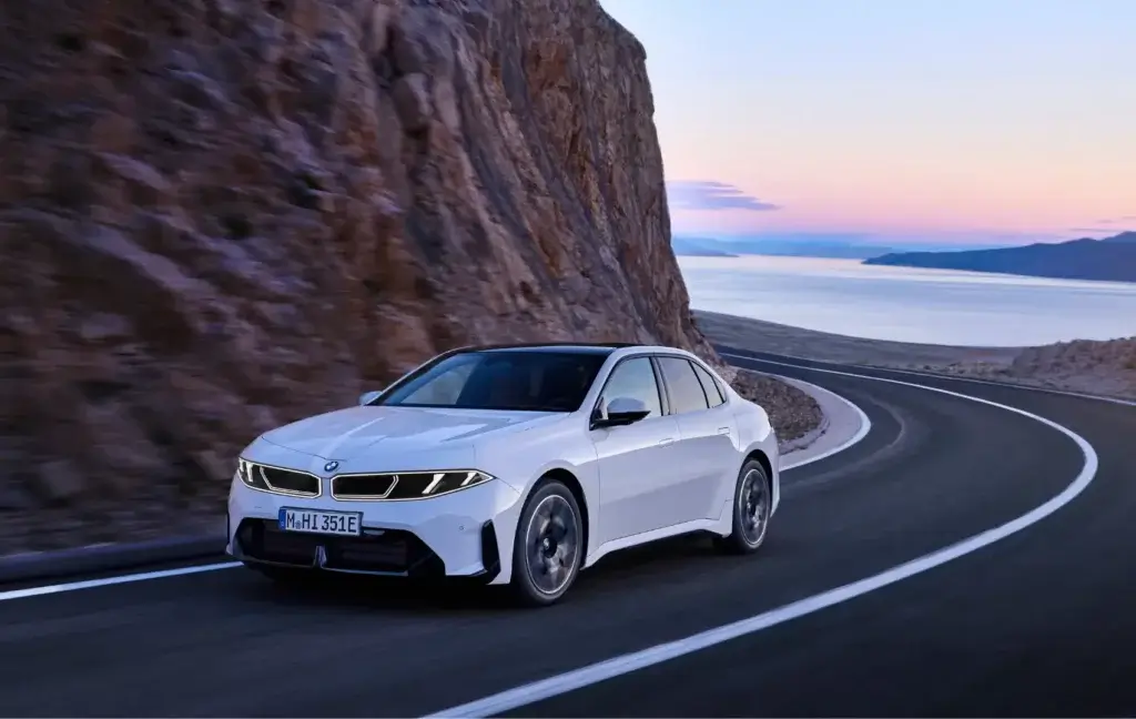 ah-praller-i3-50-xDrive-teaser Eine weiße Limousine der „Die neue Klasse“ BMW iX3 fährt bei Sonnenuntergang auf einer kurvenreichen Küstenstraße entlang felsiger Klippen, im Hintergrund sind ruhiges Wasser und ferne Hügel zu sehen. - AH Praller