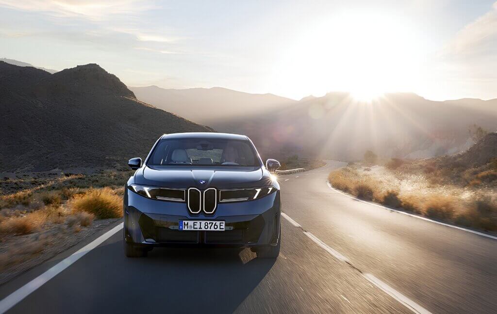 ah-praller-iX3-40-aktionsangebot Ein eleganter schwarzer BMW iX3 der „Die neue Klasse“ fährt bei Sonnenaufgang auf einer kurvenreichen Wüstenstraße, während im Hintergrund die Sonnenstrahlen über die Berge scheinen. - AH Praller